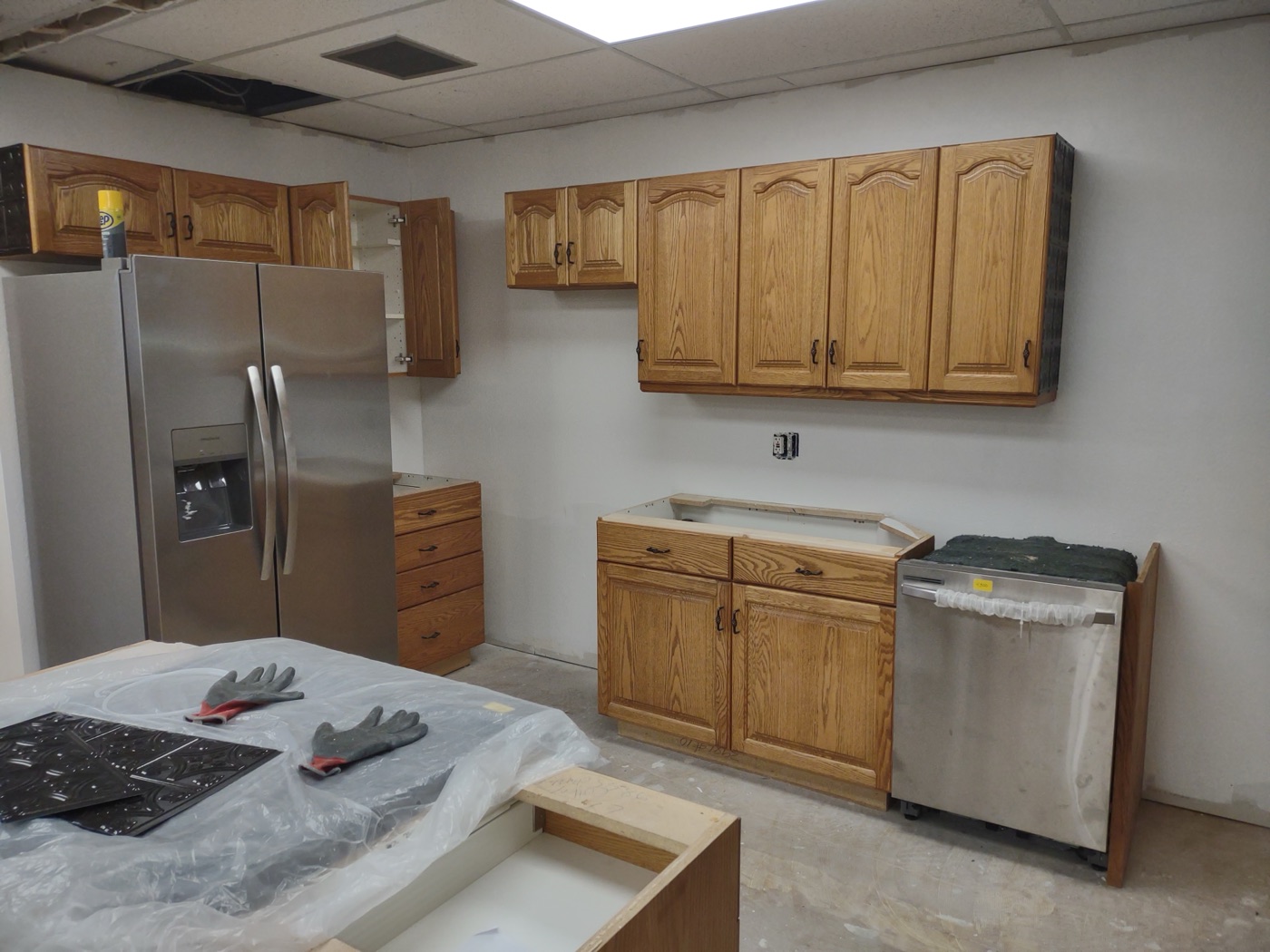 Completed kitchen install with new oak cabinets, stainless fridge, dishwasher, and cooktop wrapped in plastic